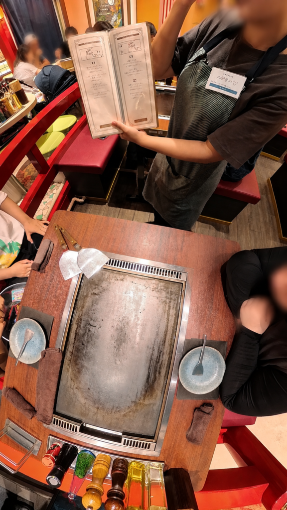店員さんがメニューを開き、もんじゃ焼きの焼き方や注文内容を説明している様子。